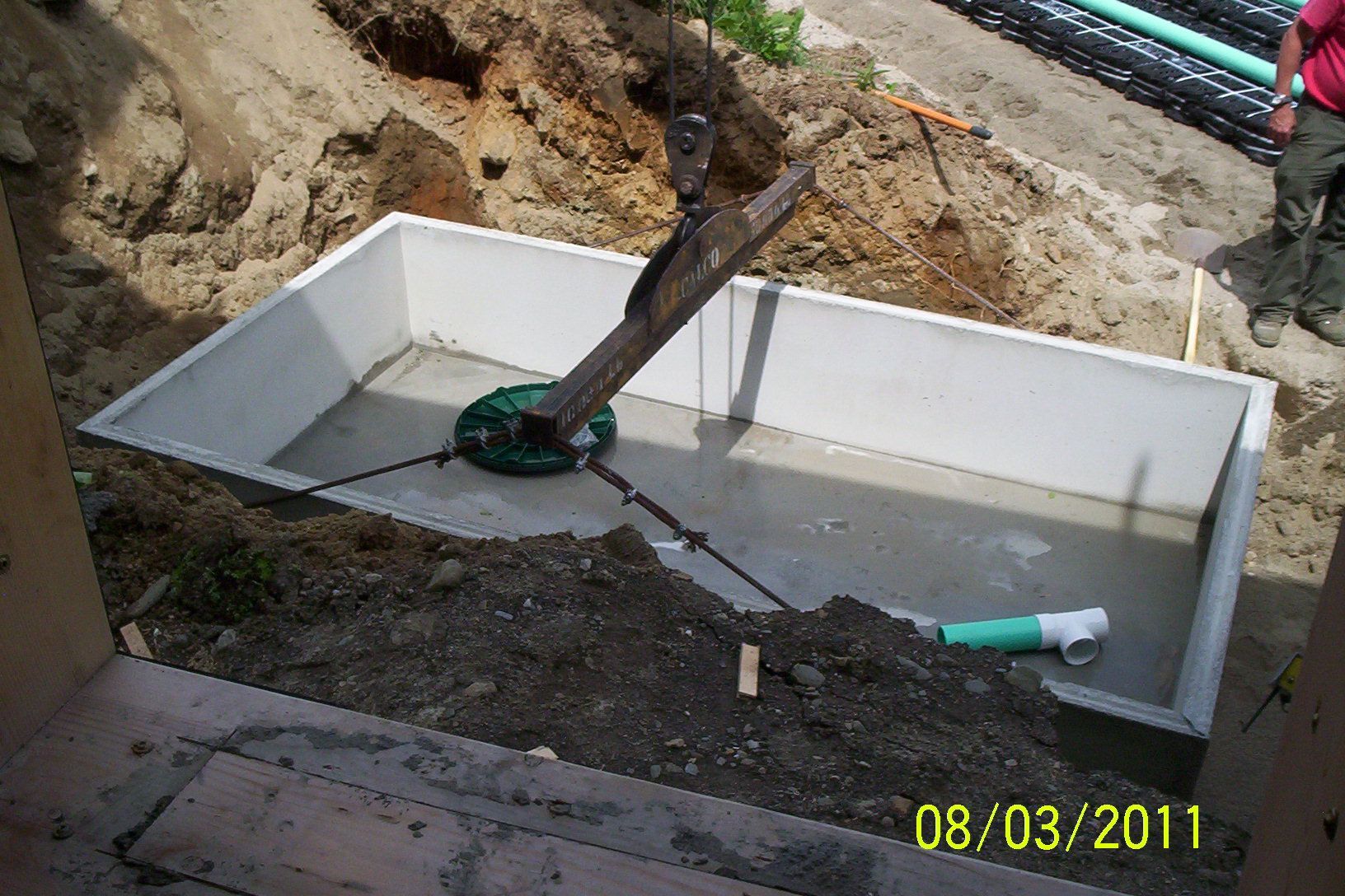 Excavator digging around a white concrete tank at a construction site, with hoses and a trench nearby.