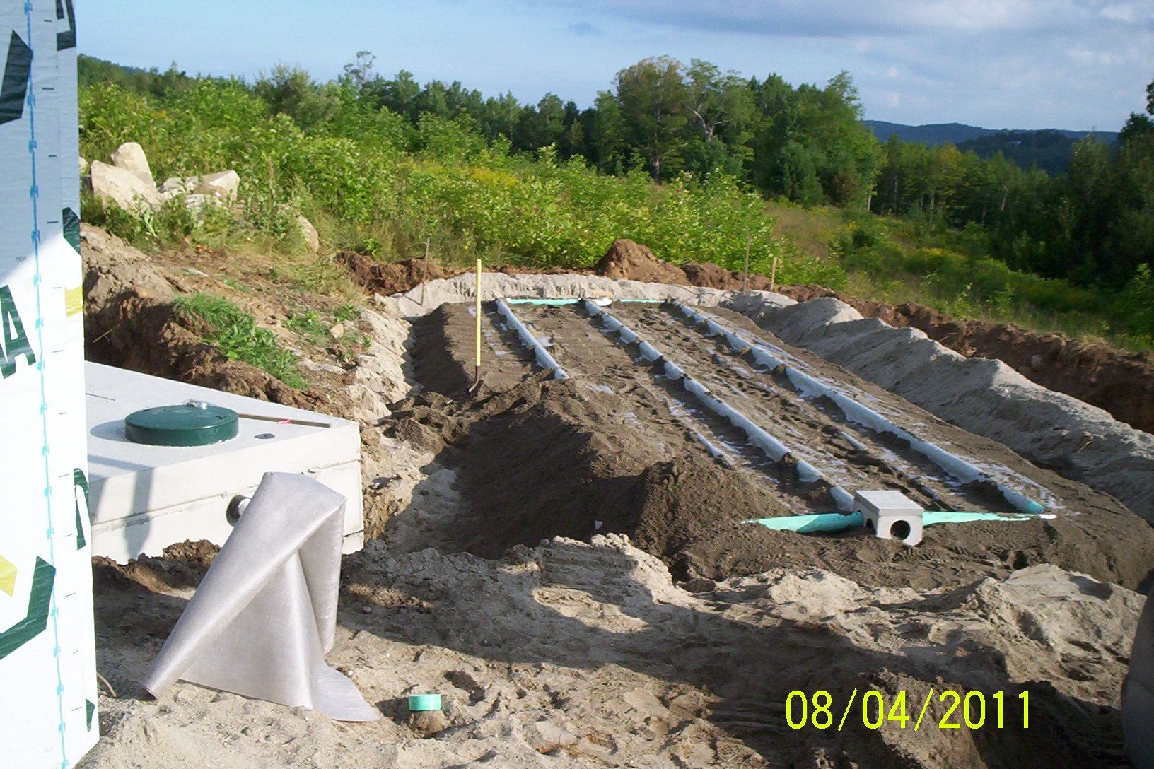 Construction site with sand trenches, white pipes, and a green hillside in the background