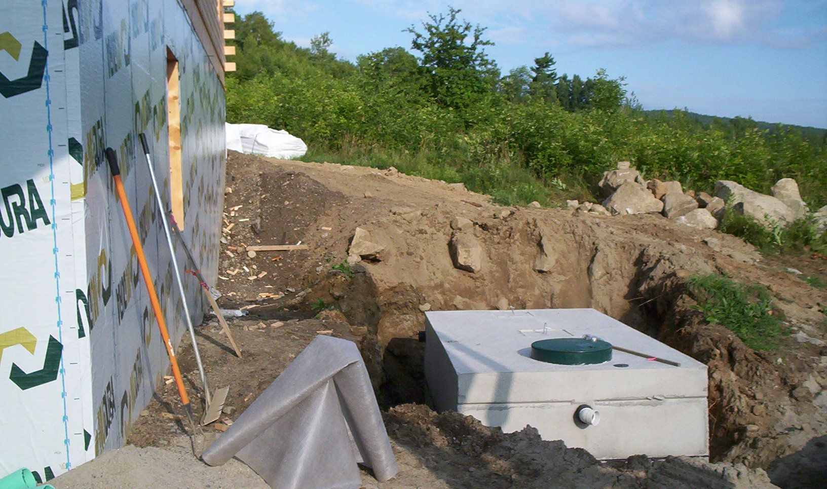 Construction site with a white concrete foundation, exposed dirt, and green brush under a blue sky