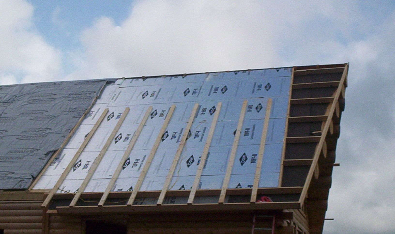 Roof under construction with blue insulation panels and wooden framing against a cloudy sky