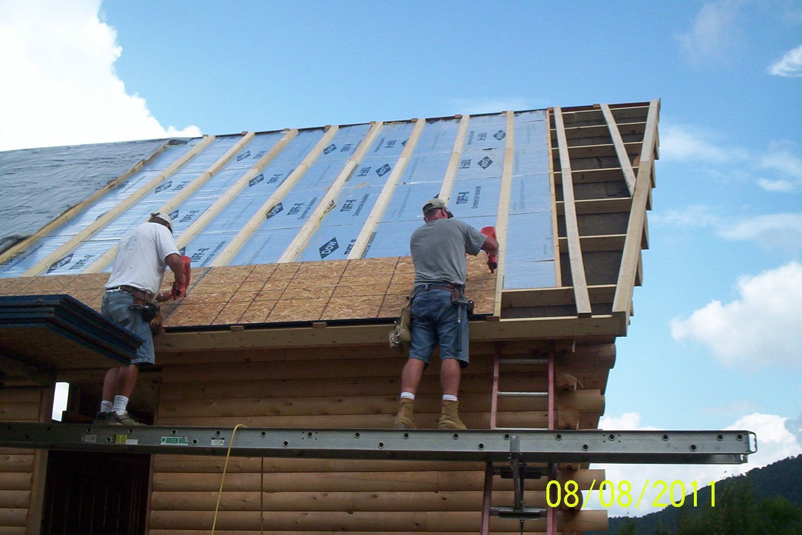 Two workers installing roof insulation and plywood on a steep wooden roof under a blue sky