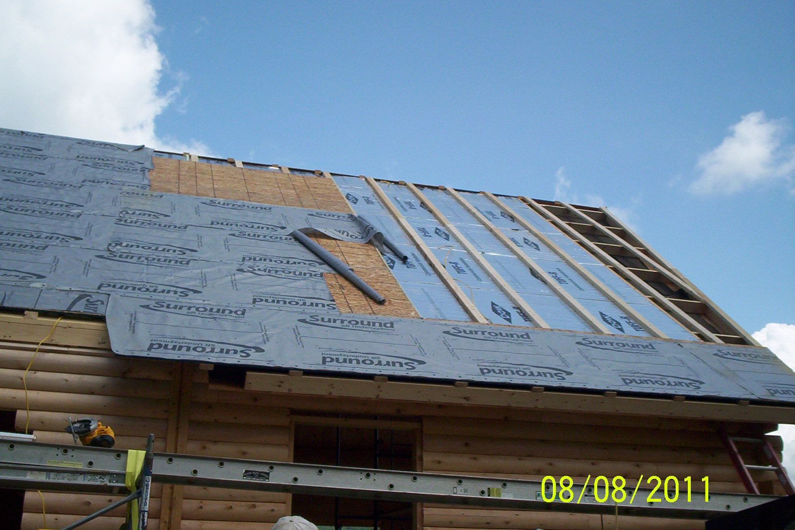 Partially shingled roof under construction with blue housewrap and exposed rafters against a cloudy sky