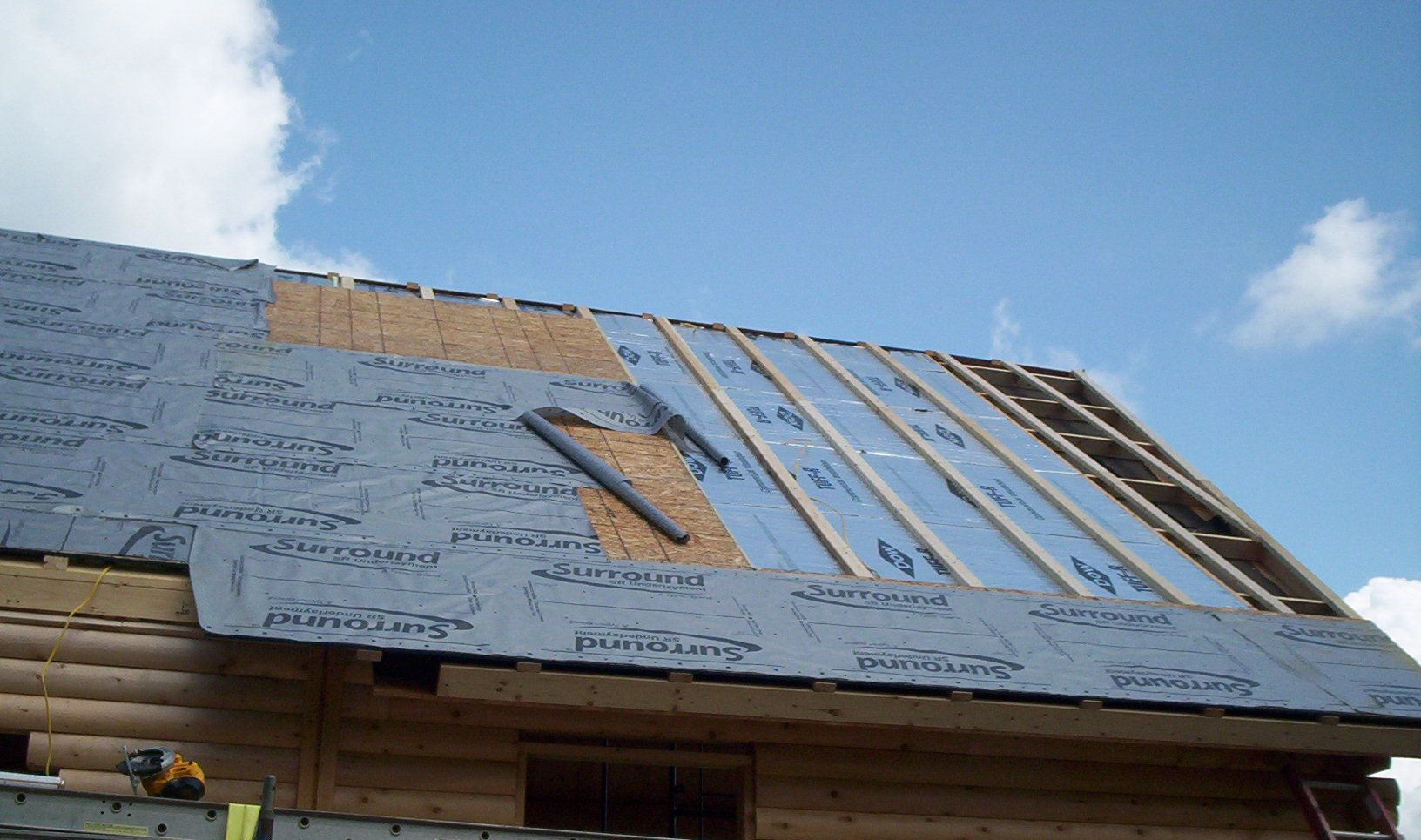 Roof under construction with blue underlayment, white framing, and open skylight area under a cloudy sky