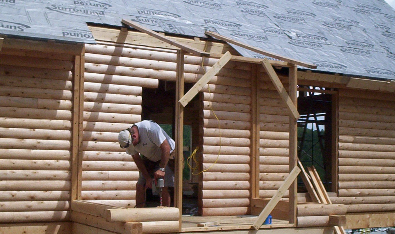 Worker building a log cabin wall, fitting timber logs beneath a shingled roof.