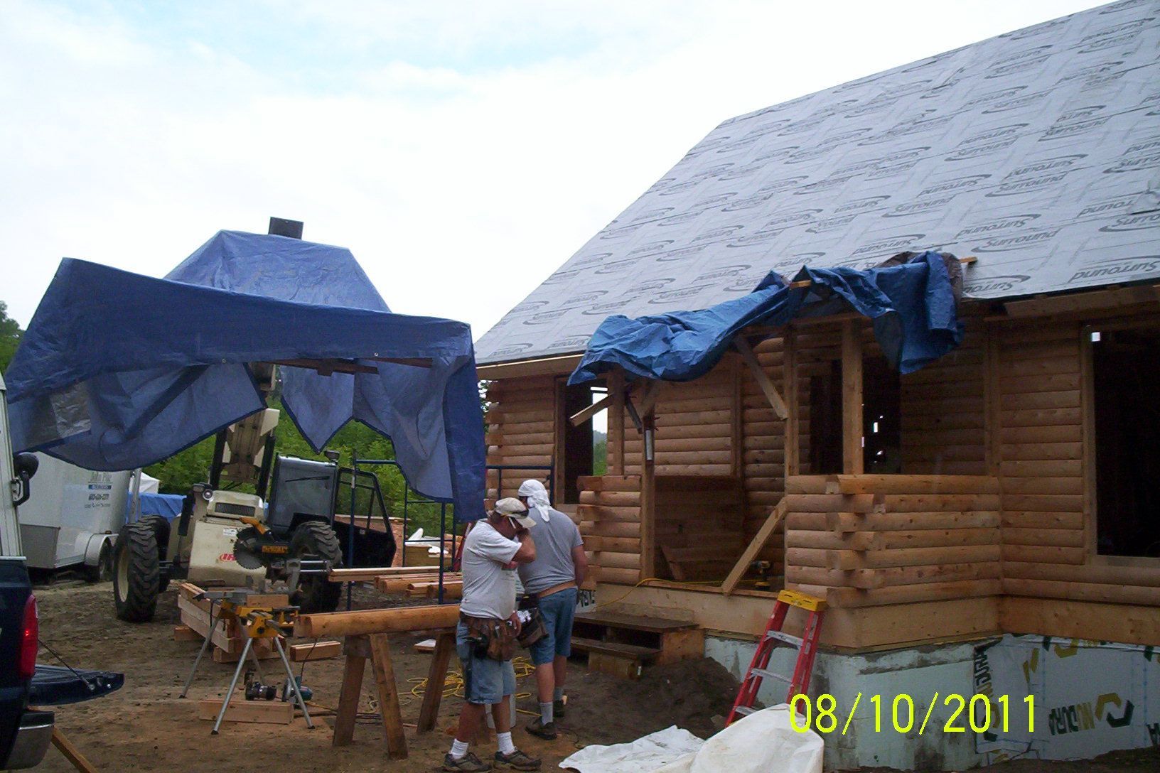 People standing by a log cabin under construction with blue tarps covering the roof and canopy.