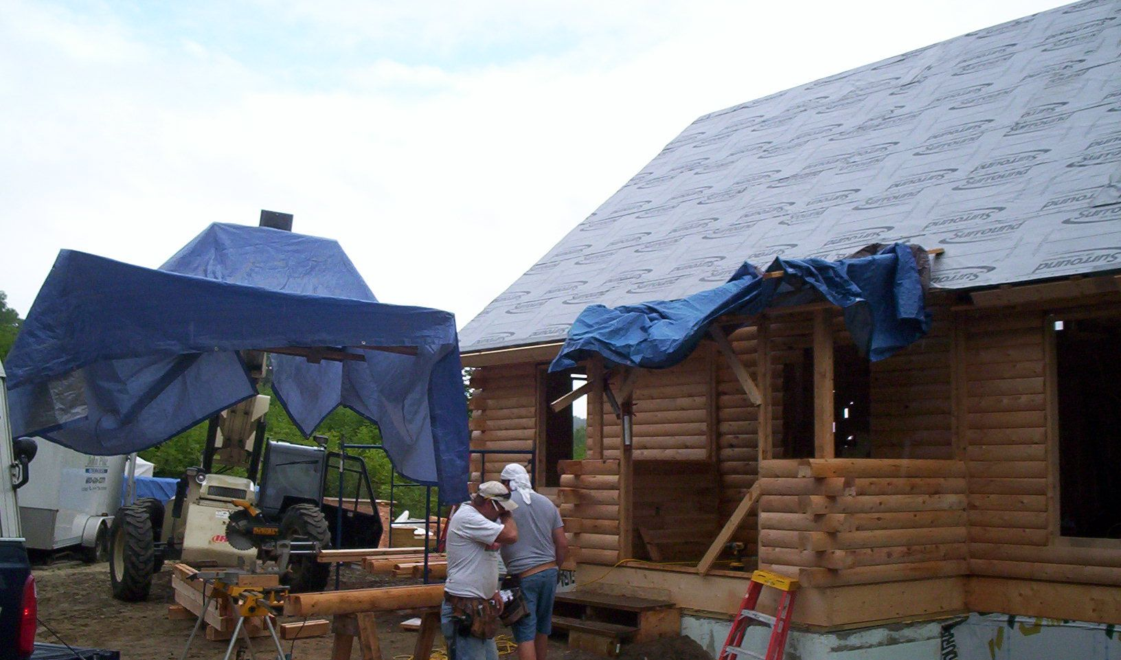 Workers constructing a log cabin with blue tarp-covered scaffolding beside a building with a steep roof