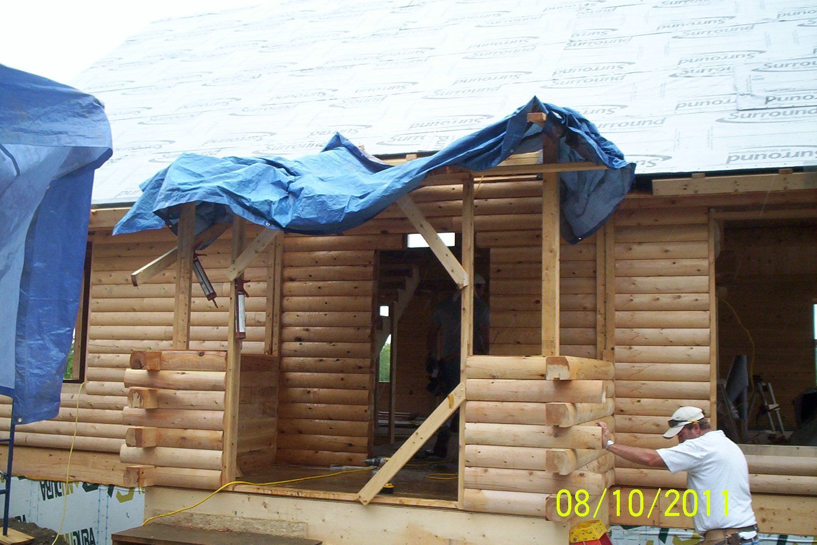 Wooden log cabin under construction with a blue tarp covering the roof and entrance supports exposed
