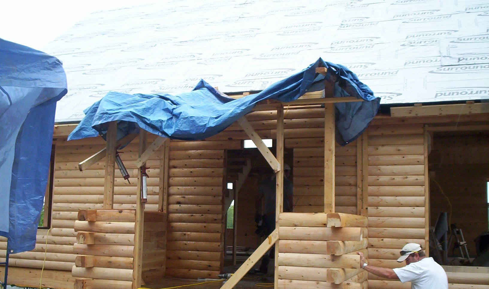Log cabin under construction with a blue tarp draped over the roof and entrance porch