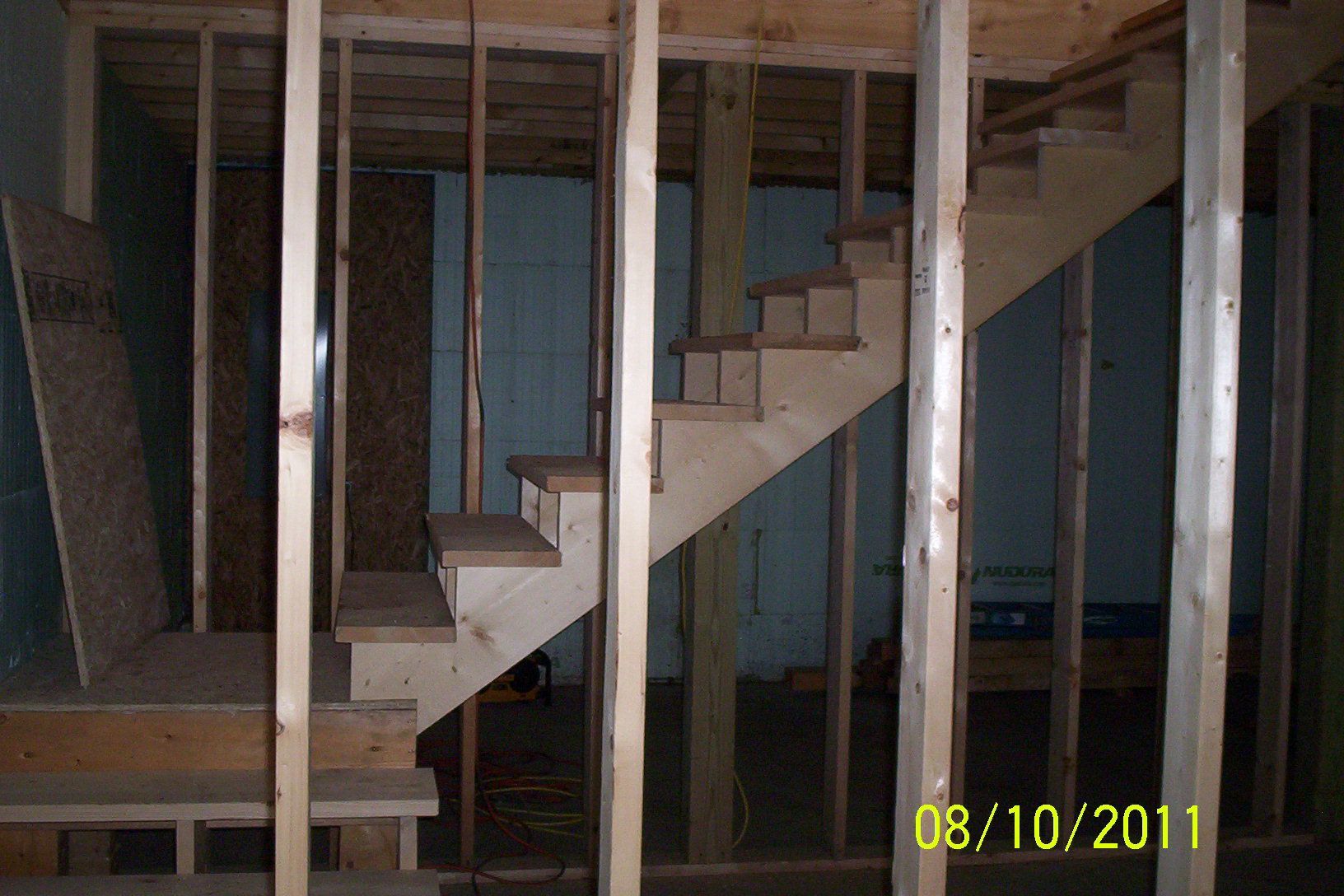 Unfinished wooden staircase framed by studs and blue wall panels in a construction site.