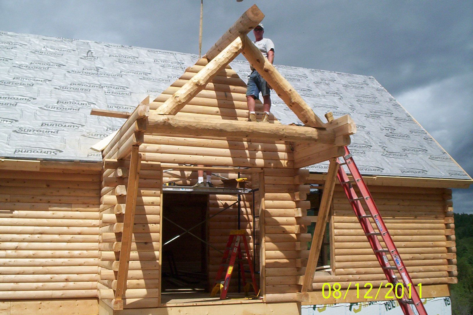 Workers building a log cabin roof, with ladders and exposed wooden beams under a cloudy sky