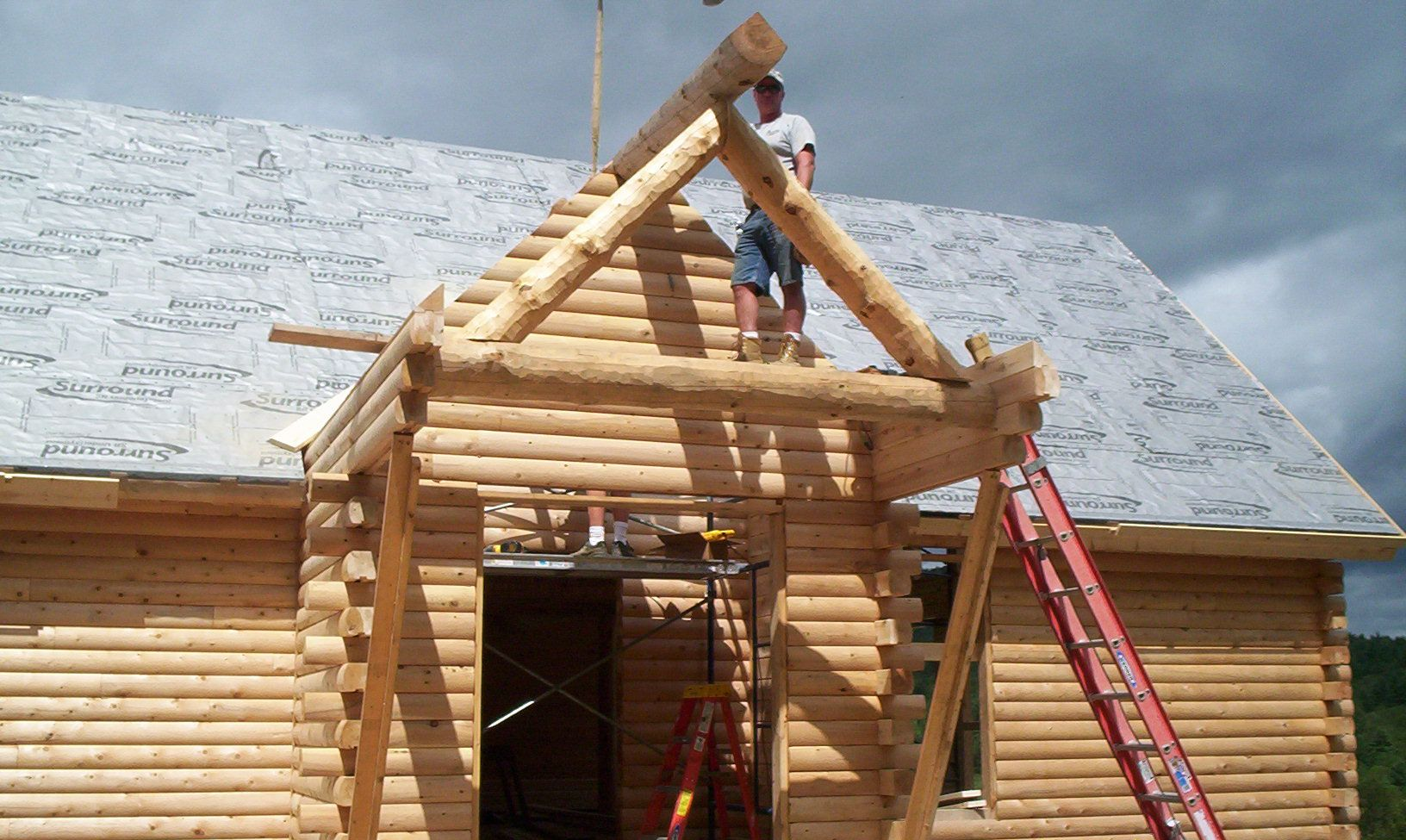Workers constructing a log cabin roof with ladders and exposed rafters under a cloudy sky
