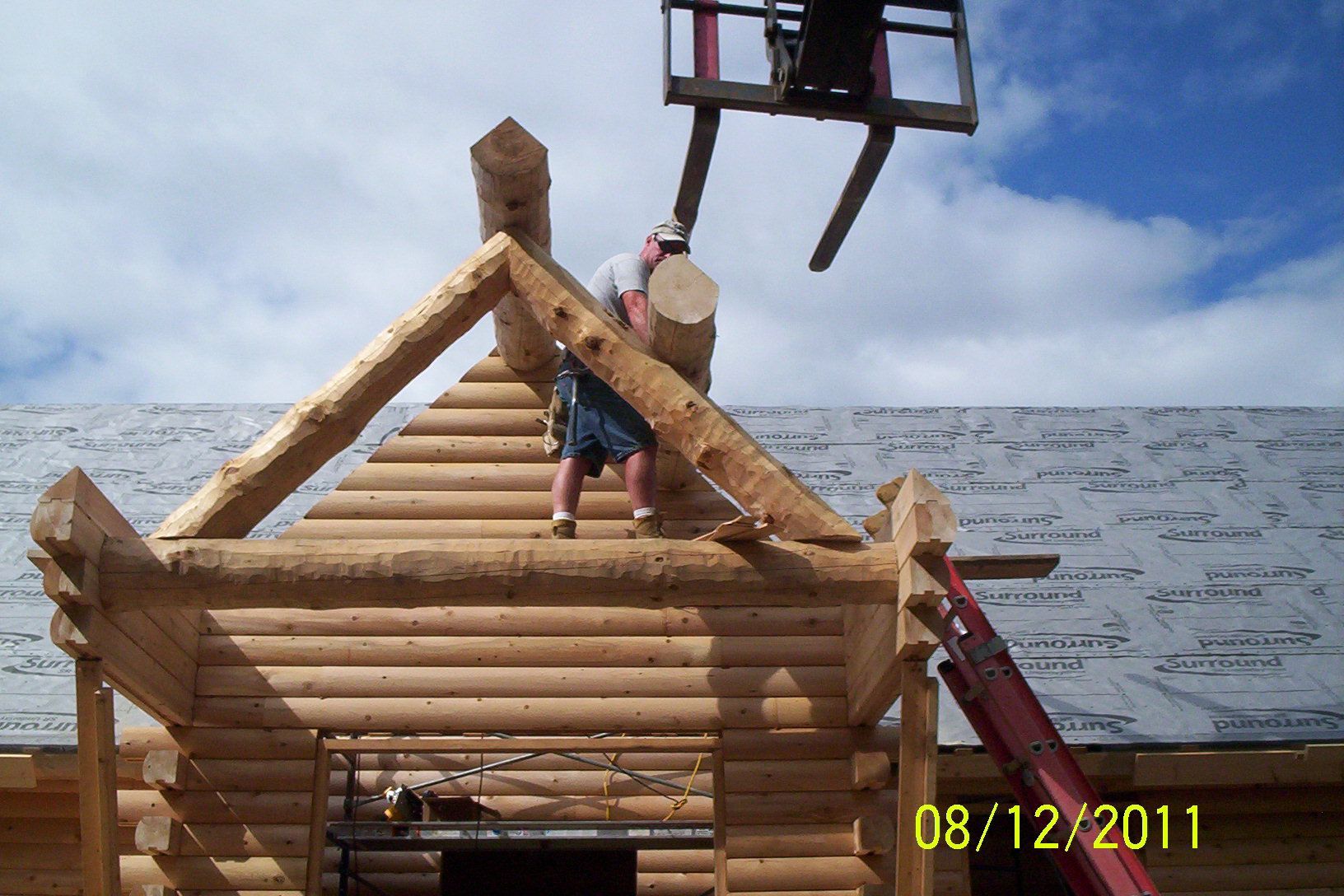Two workers on a log cabin roof lifting a beam under a blue sky