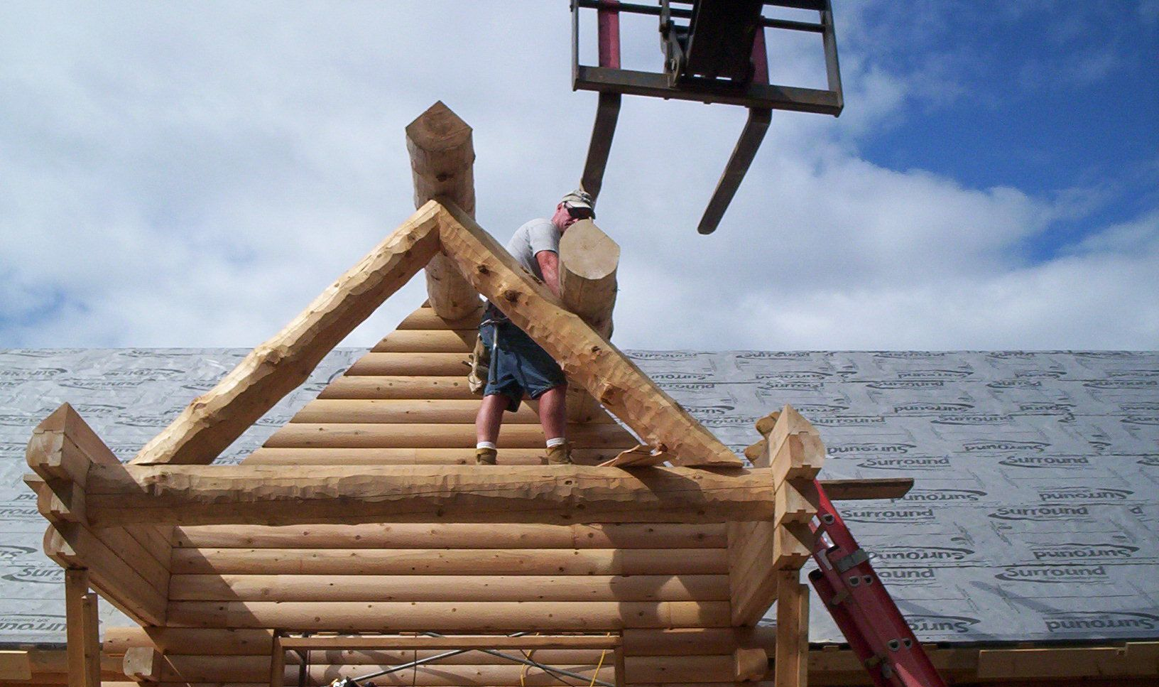Worker on a log cabin roof under construction, lifted by a crane against a blue sky.