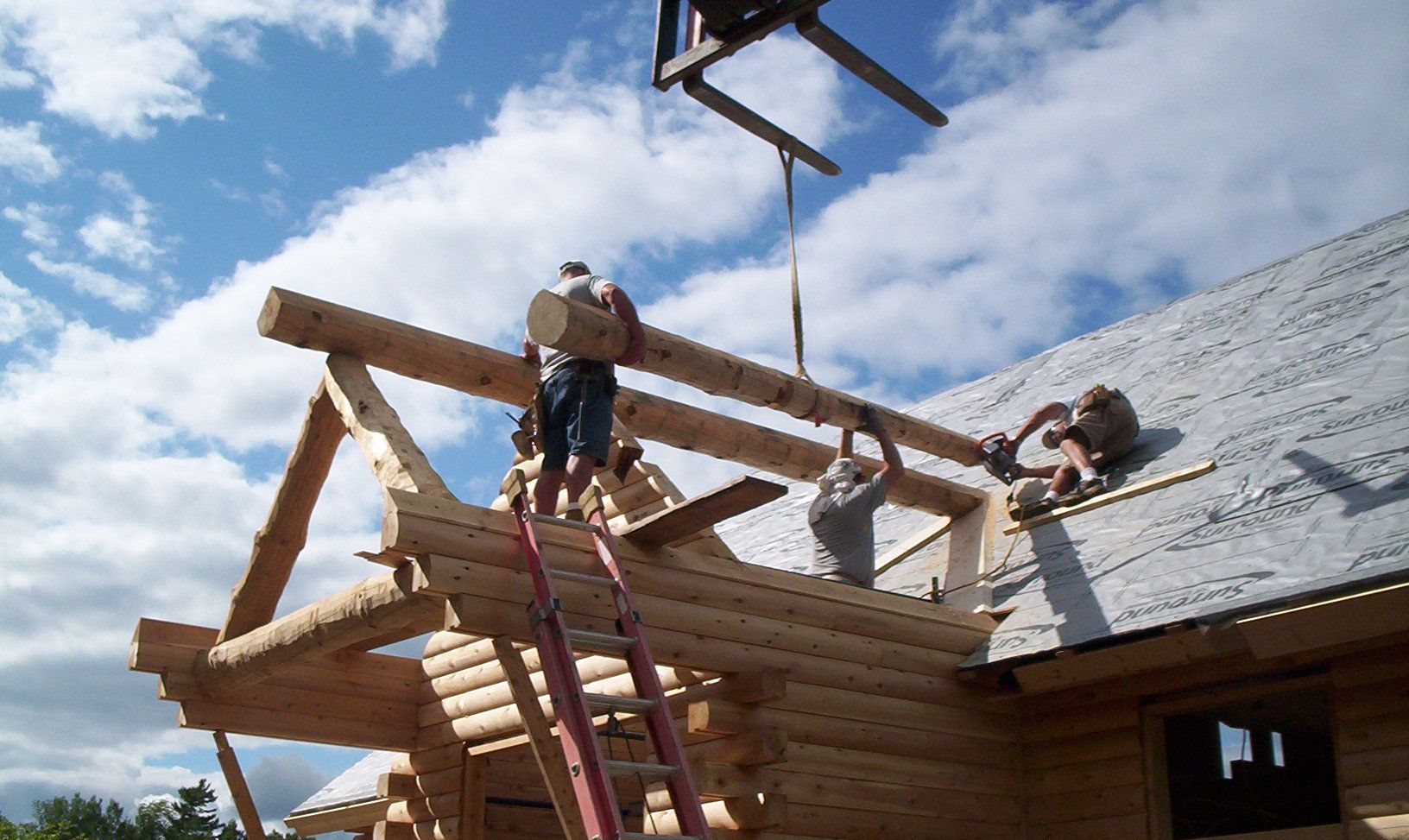Workers installing wooden roof beams on a log cabin under a partly cloudy sky