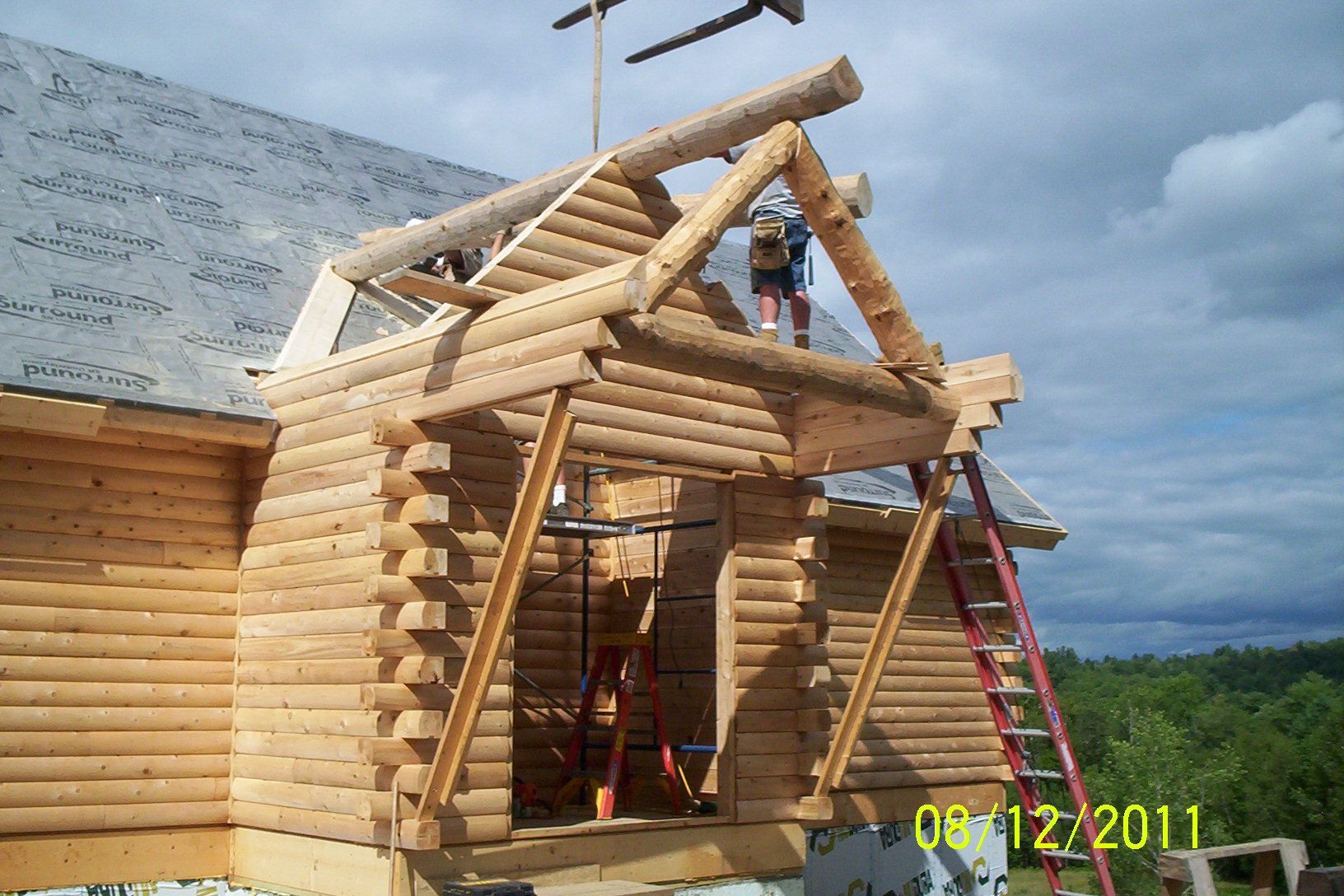Log cabin under construction with exposed log rafters and roof framing, date stamp visible at bottom right