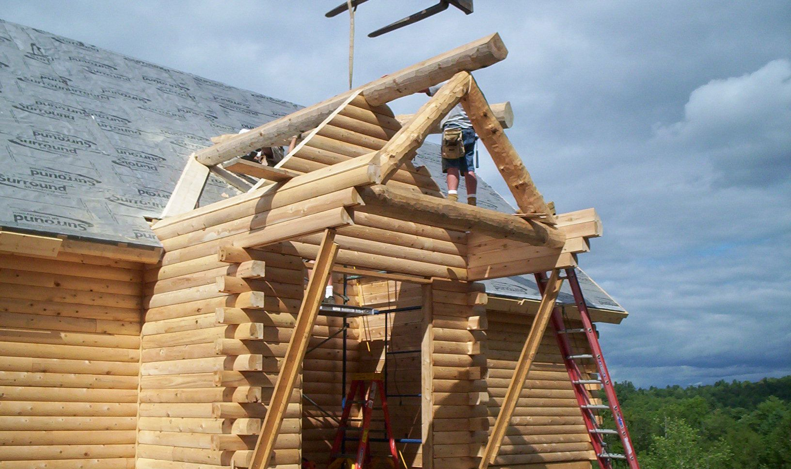 Log cabin under construction with log rafters, roof framing, and two ladders against a cloudy sky.