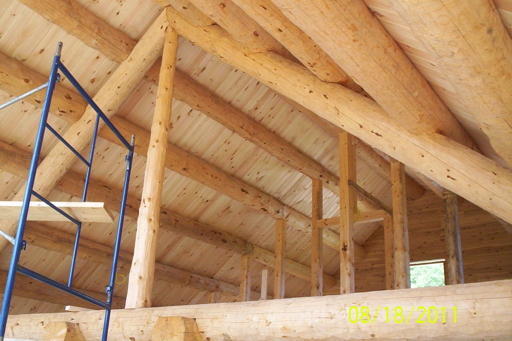 Interior of a wooden house under construction, with blue scaffolding and exposed roof beams.