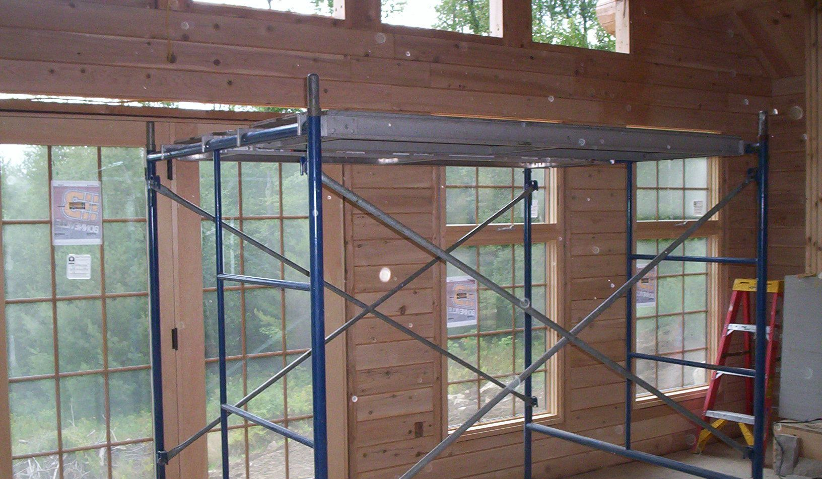 Scaffolding inside a wood-framed building with large windows and a red ladder on the right