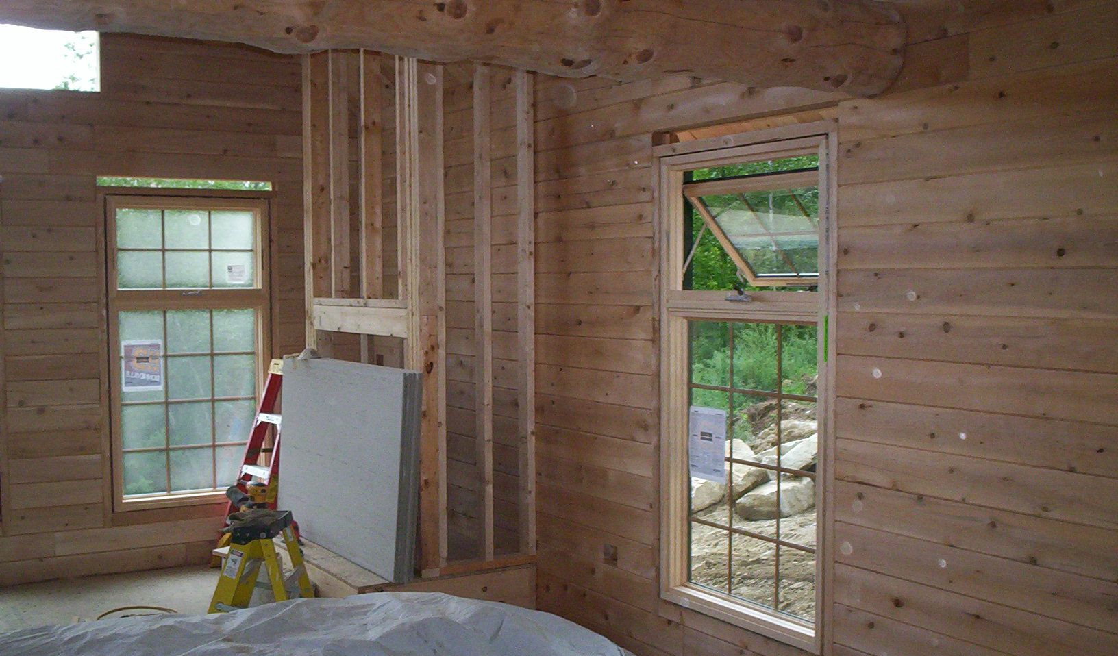 Unfinished room with exposed wood studs, two windows, and a ladder leaning near a drywall panel.