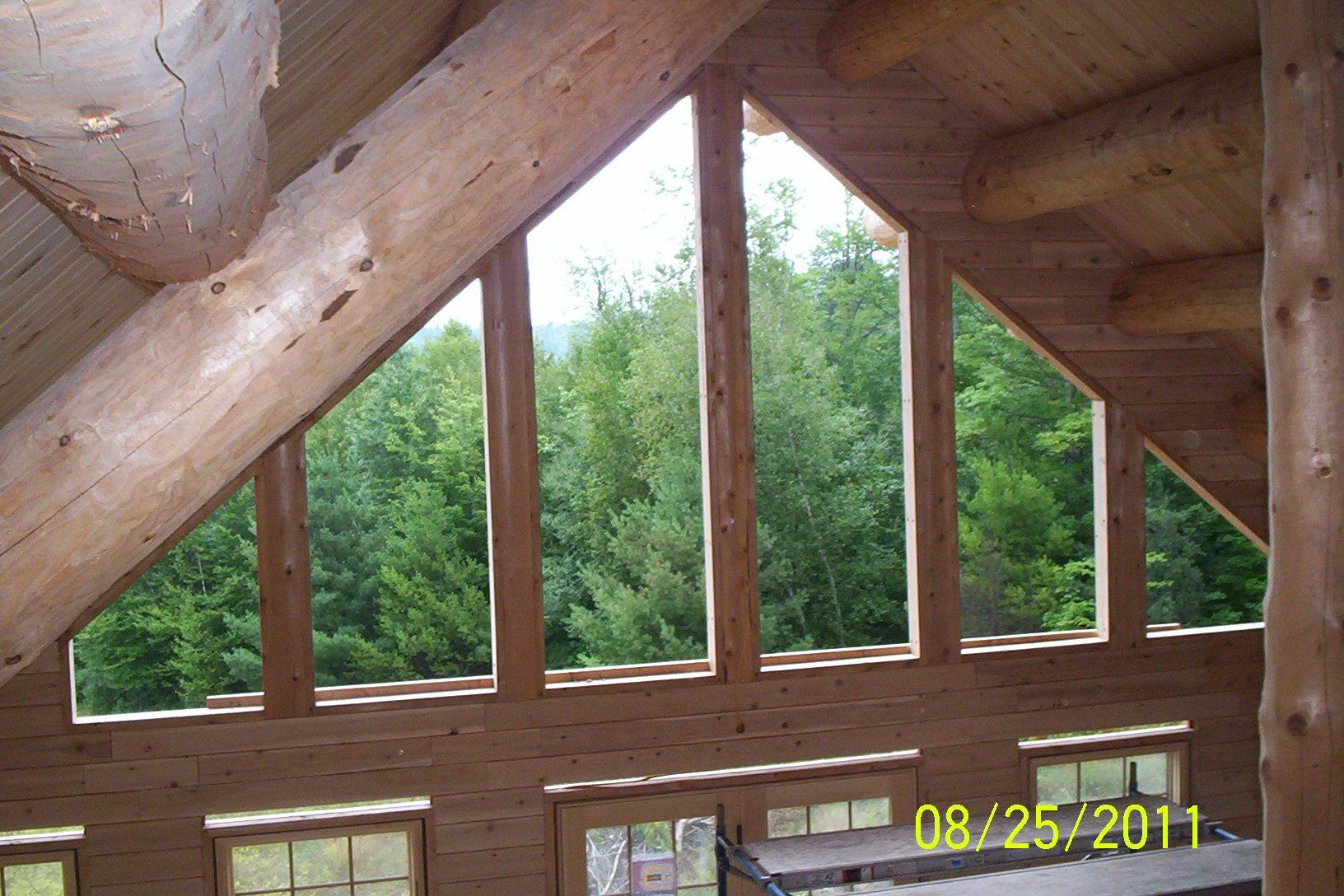 Wooden cabin interior with large triangular windows overlooking green trees.