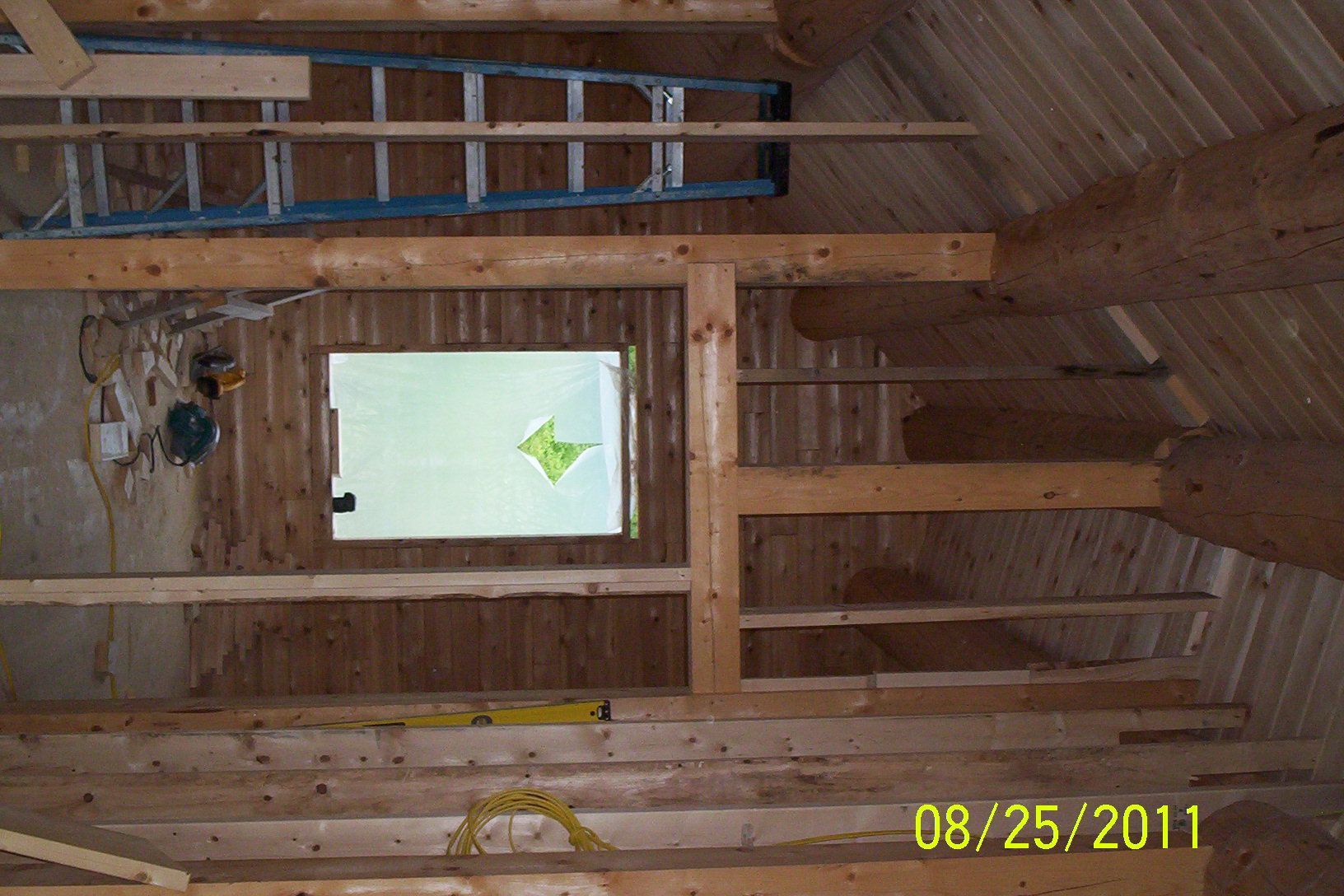 Attic interior under construction with exposed wood framing, a window, and a ladder on the rafters.
