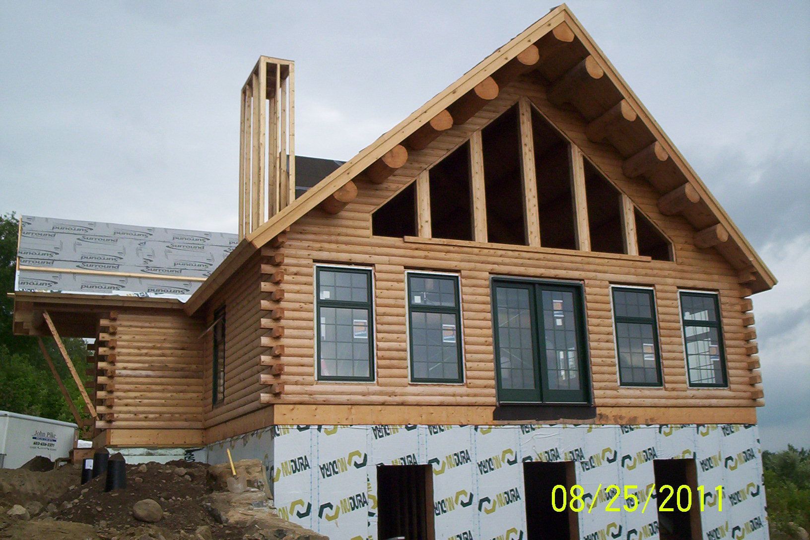 Partially finished log house with gabled roof and blue windows on a construction site