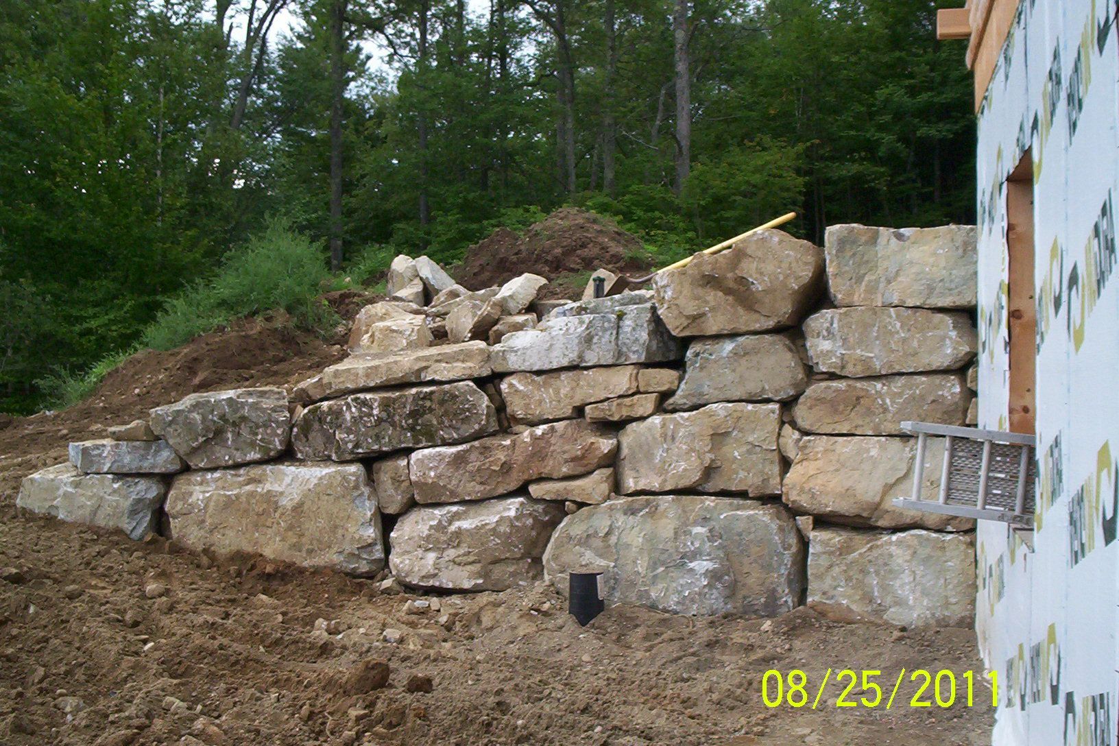 Stone retaining wall under construction on a dirt slope beside a wooded area.