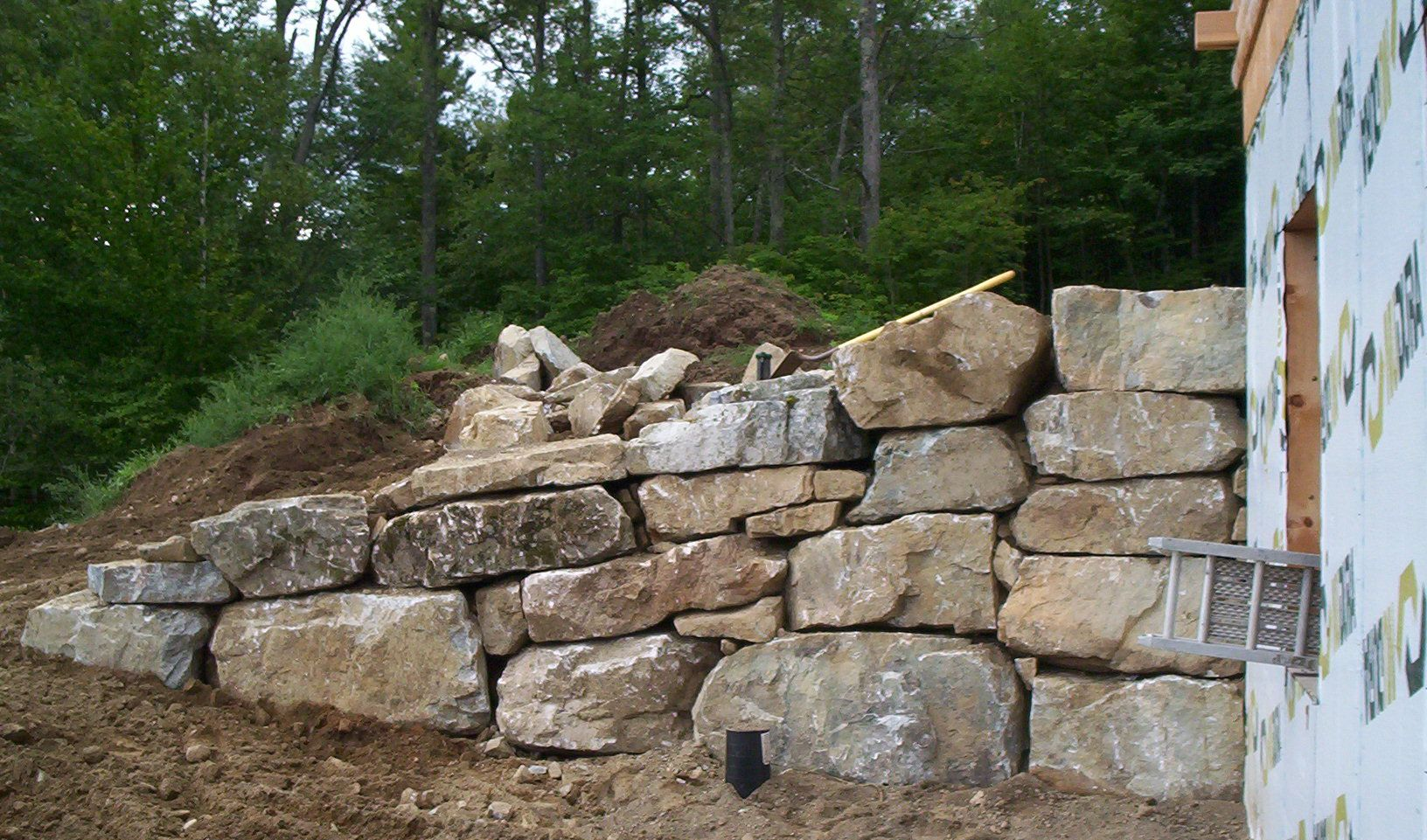 Stone retaining wall built into a dirt slope beside a white building, with trees in the background
