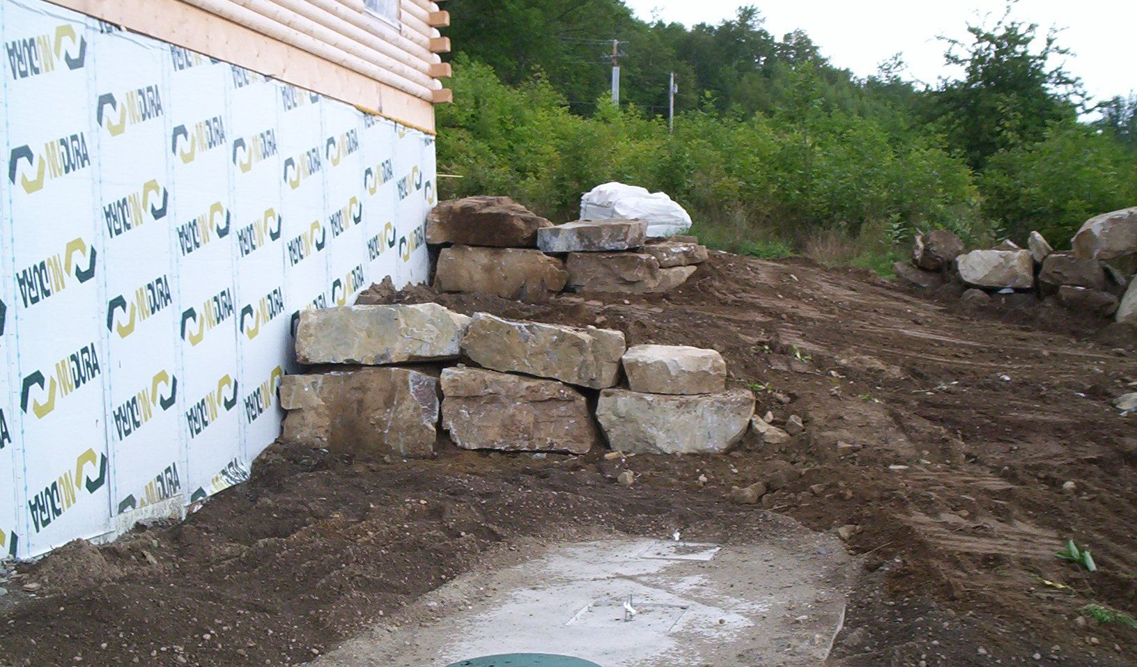 Rock retaining wall beside a house under construction on a dirt slope with scattered boulders.
