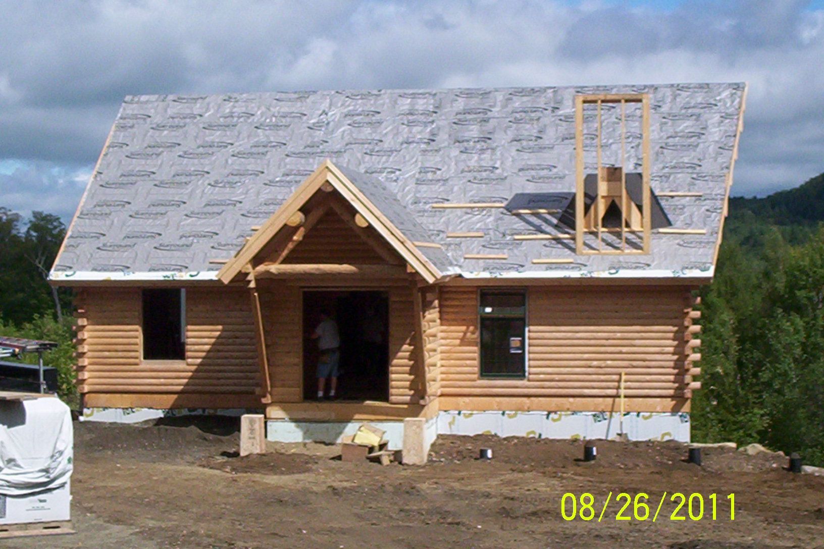 Small log cabin under construction with a gray roof and open doorway, on a dirt lot under cloudy sky.