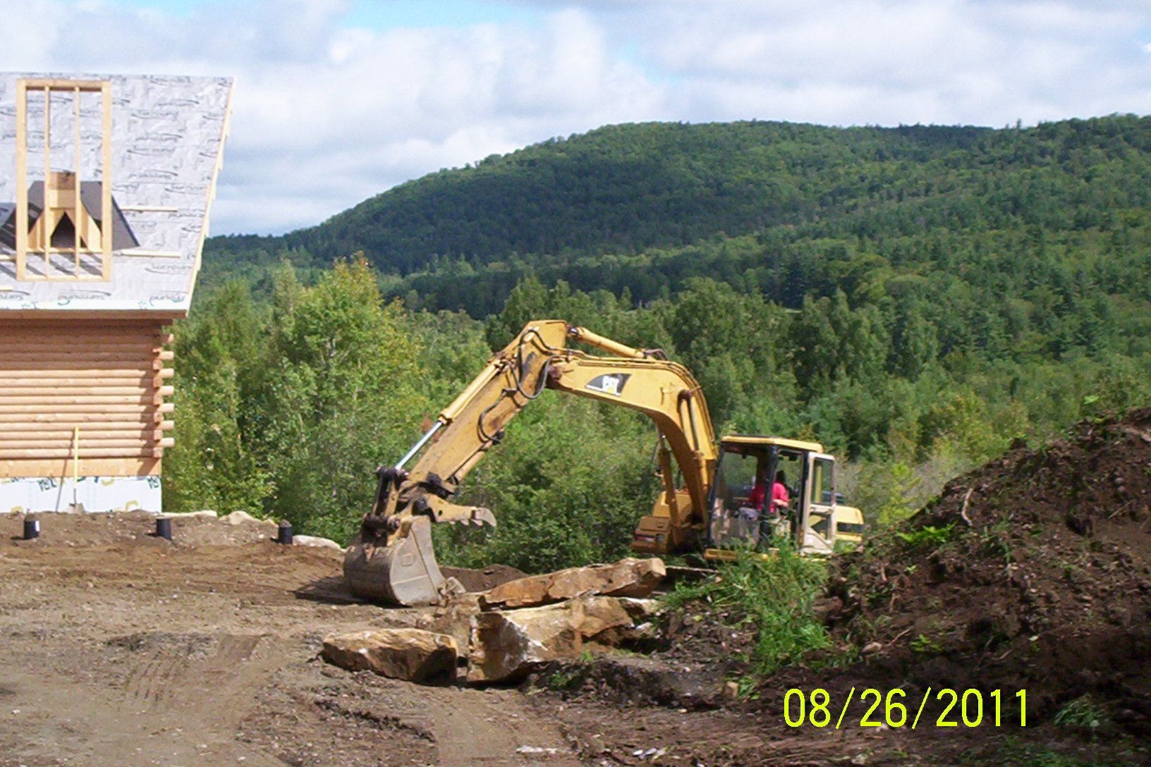 Excavator moving dirt at a construction site with a house frame and forested hills in the background.