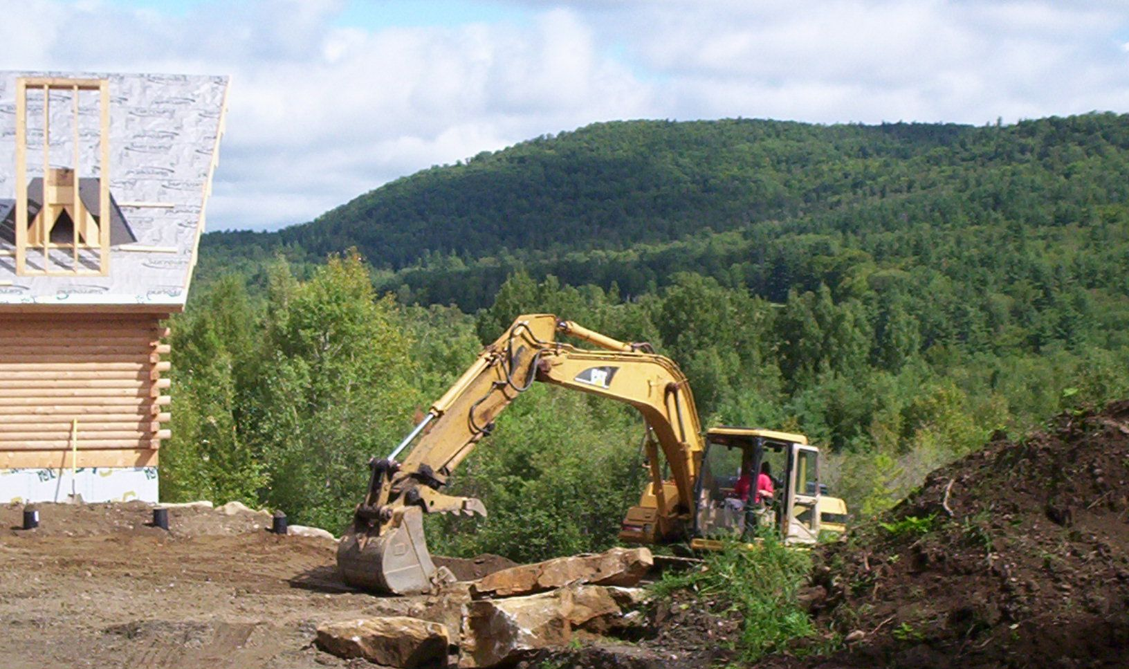 Yellow excavator at a construction site beside a partially built house and wooded hillside.