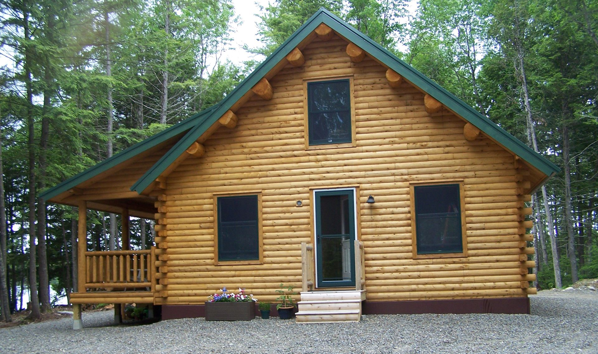 Log cabin with green roof trim and front porch, surrounded by trees on a gravel lot