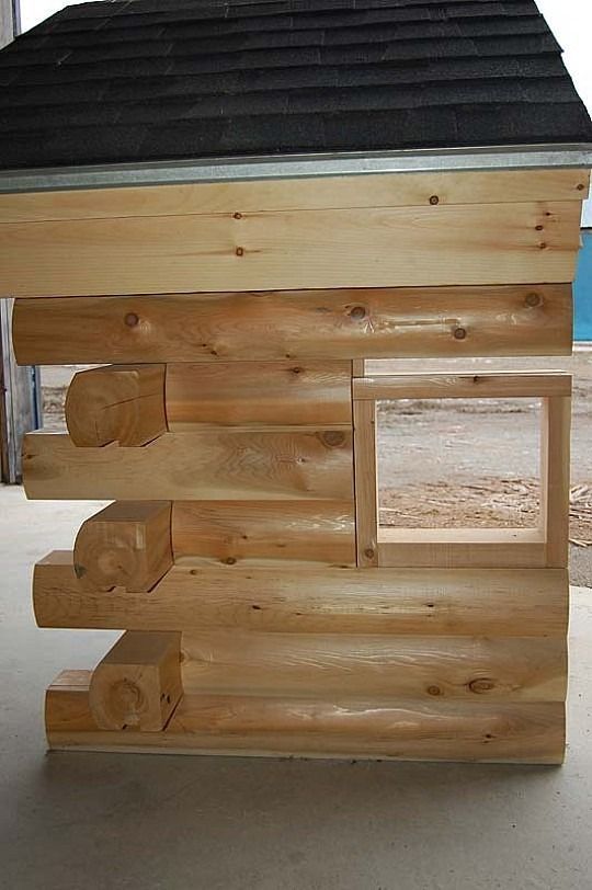Wooden log cabin playhouse with a black roof, small windows, and a side door opening.
