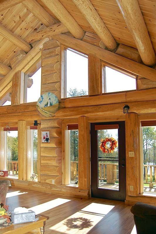 Sunlit log cabin living room with vaulted wooden ceiling, large windows, and a glass door with wreath