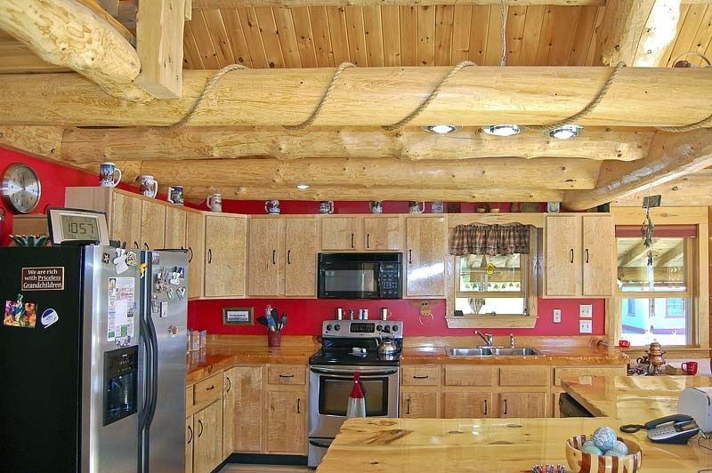 Rustic kitchen with wood cabinets, stainless appliances, and red backsplash under log beams