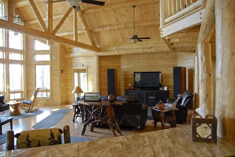 Spacious log cabin living room with large windows, wood beams, and rustic furniture around a TV