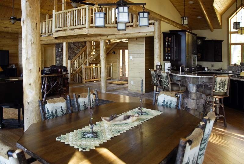 Rustic cabin great room with wood beams, stone fireplace, and dining table overlooking a loft and large windows