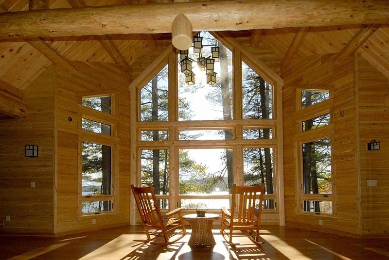 Wooden cabin interior with tall windows, chairs, and sunlight overlooking a lake