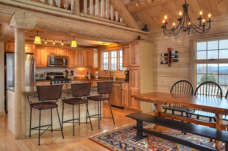 Rustic cabin kitchen and dining area with wood beams, bar stools, and a long wooden table under a chandelier