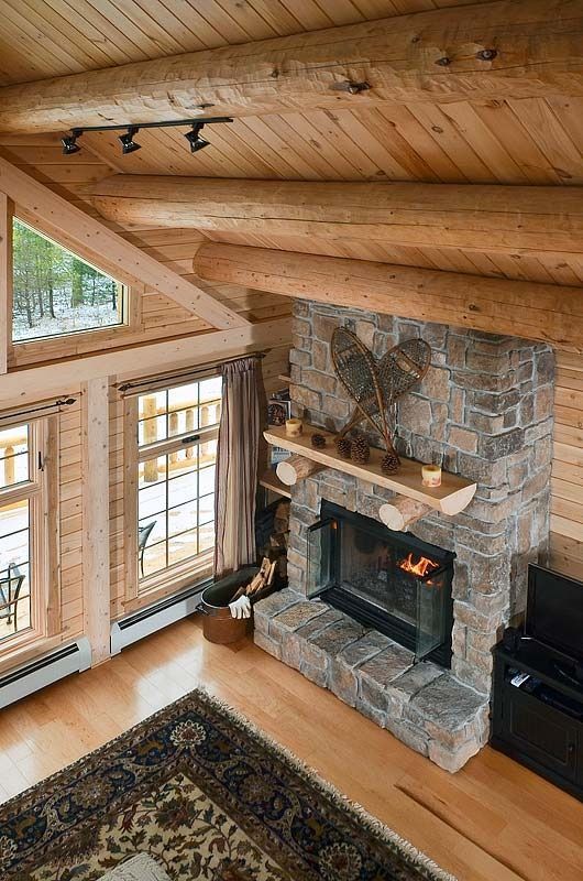 Rustic living room with stone fireplace, wood ceiling, and tall windows overlooking a rug and hardwood floor