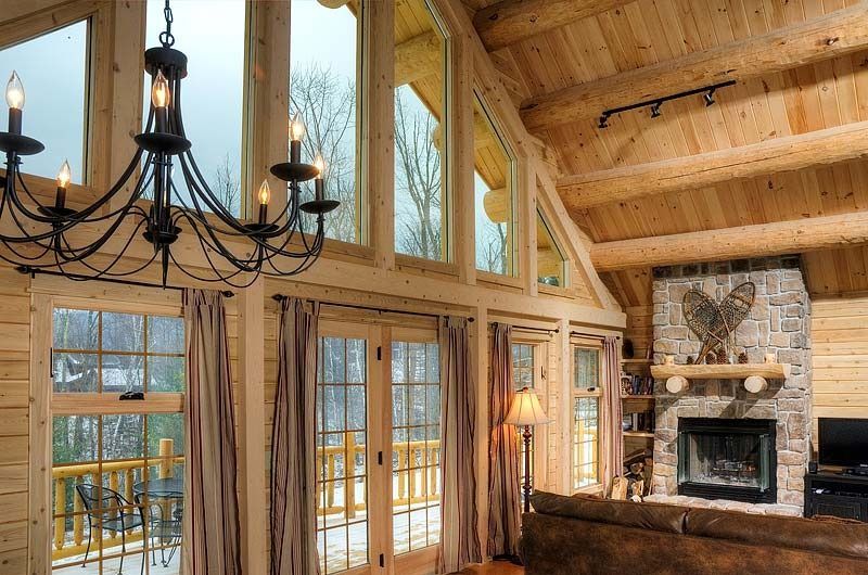 Cozy log cabin living room with stone fireplace, large windows, wood beams, and black chandelier