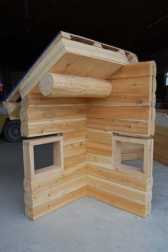 Wooden log playhouse corner with two small windows and a log roof, indoors on a concrete floor