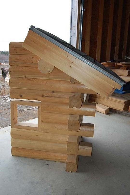 Wooden log cabin model with a slanted roof on a concrete floor, near a wall and doorway.
