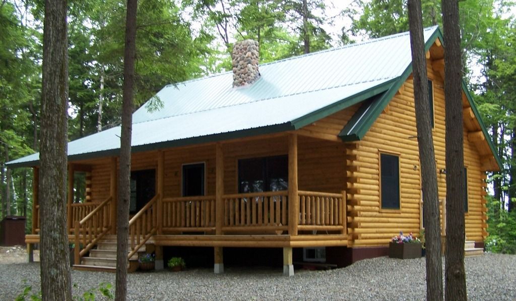 Wooden cabin with a green roof in a wooded area, with a front porch and gravel yard