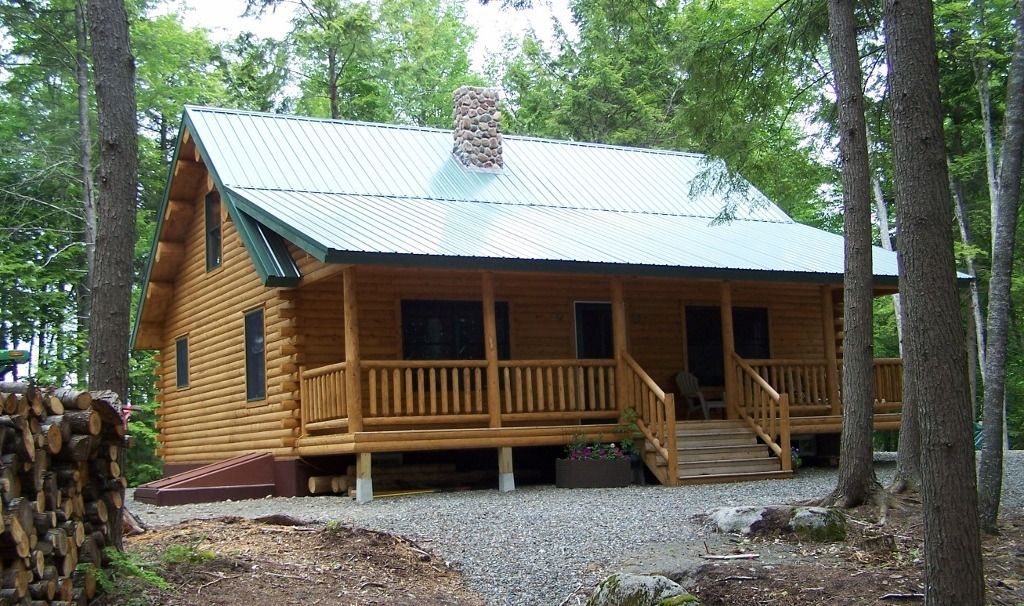 Wooden cabin with a covered porch in a wooded area, surrounded by trees and gravel.