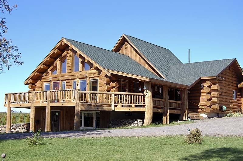Large log cabin with wooden decks and a gray roof on a grassy lot under a blue sky