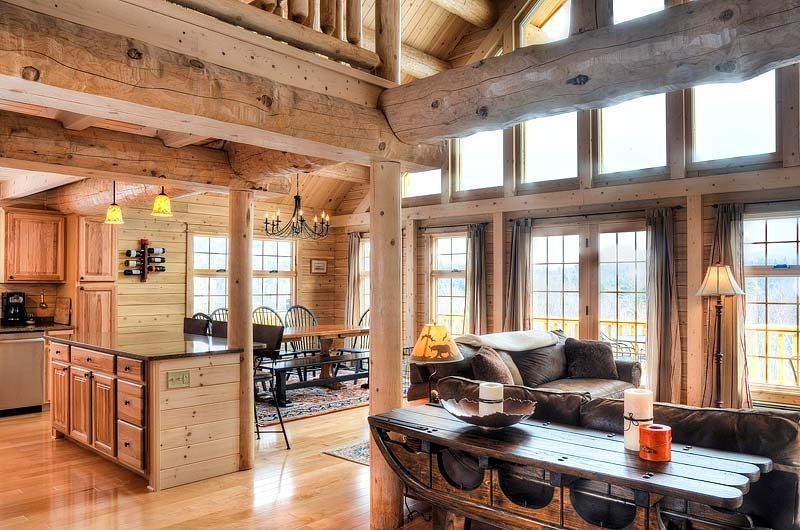 Cozy rustic cabin kitchen and living area with wooden beams, large windows, and a stone fireplace