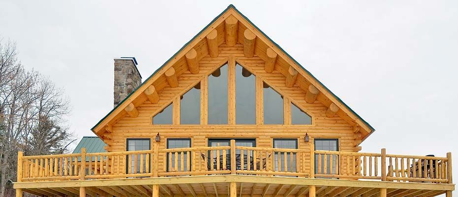 Wooden cabin with a large front deck, stone chimney, and tall peaked roof in a snowy setting