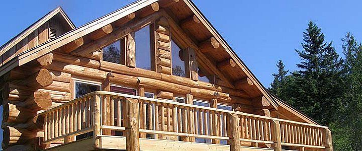 Log cabin with a wooden deck under a blue sky, surrounded by evergreen trees